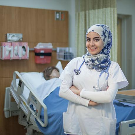 nursing student smiles while crossing arms st和ing in front of hospital bed in sim lab